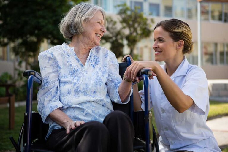 Two caregivers chatting with an elderly resident in a bright, tidy common room with comfortable seating.